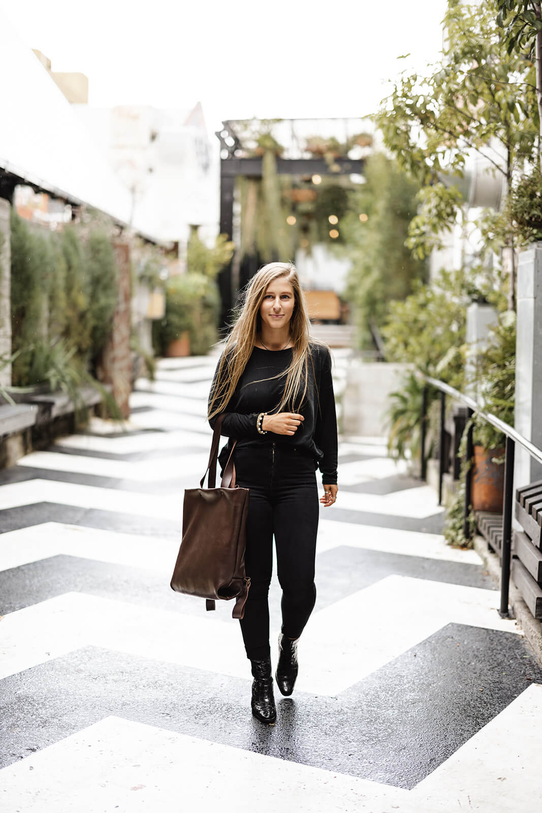 Blonde woman in black clothes walking along black and white zigzag ground surrounded by greenery. She is wearing a brown leather tote on the inside of her elbow. The tote is the Chocolate Brown Leather Backpack & Tote by Ella Jackson