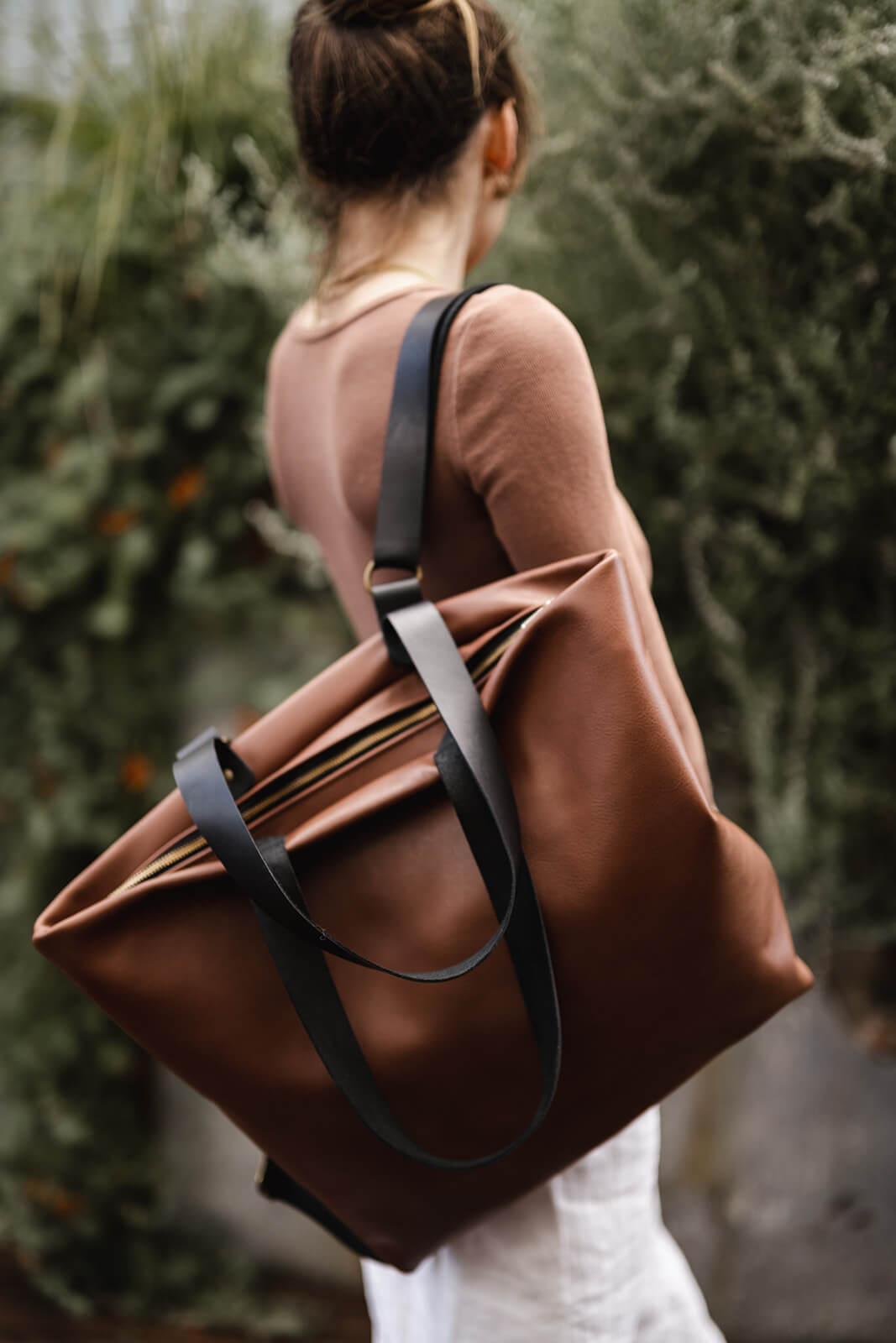Woman with back to camera with dark hair up and brown top and white skirt standing in front of greenery. She is modelling the Tan Leather Backpack & Tote with Black straps by Ella Jackson