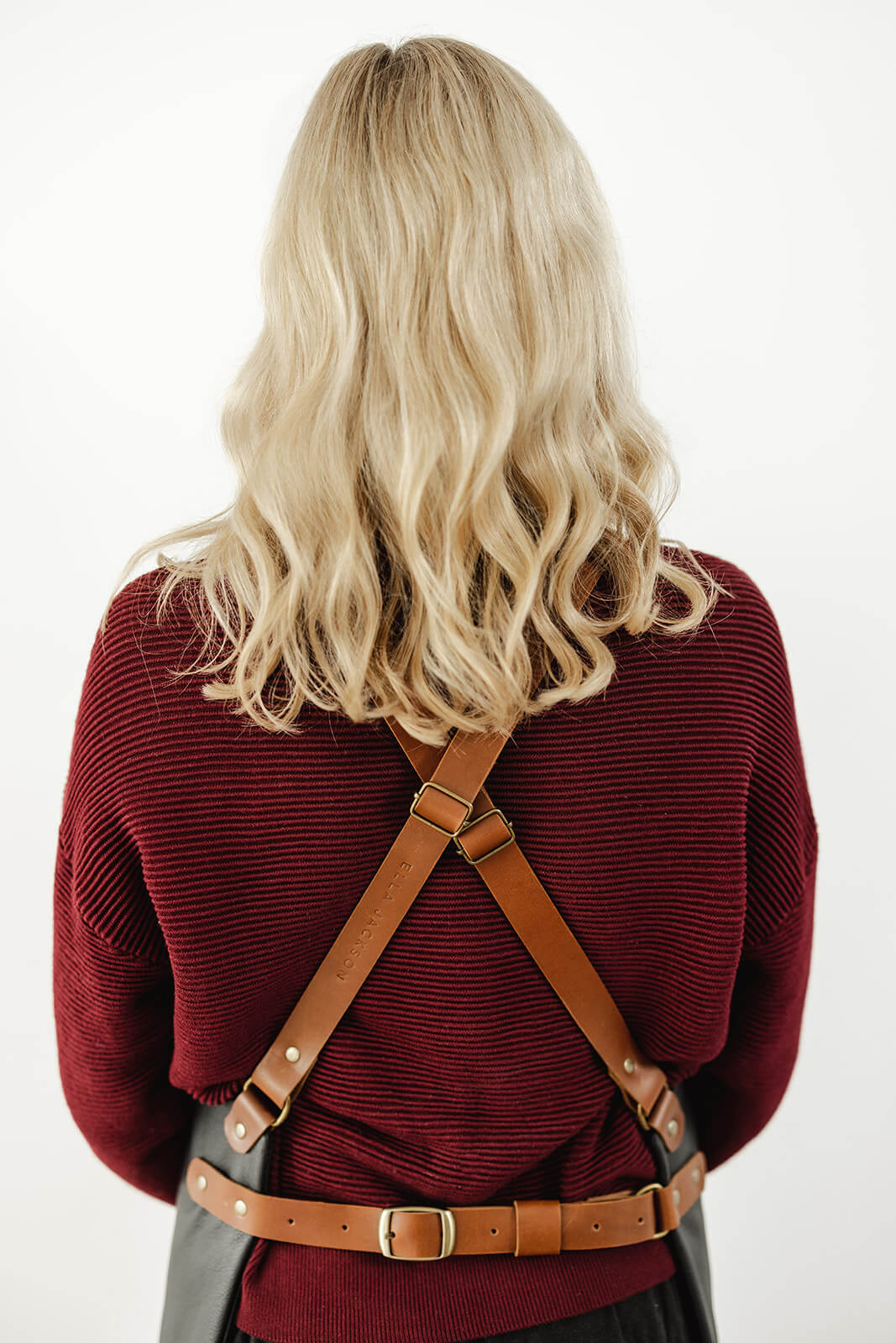 Back view of woman with blonde wavy hair and maroon jumper wearing a leather apron. The pic shows the strap configuration from the back...tan leather crisscross shoulder straps, a waist strap and antique hardware