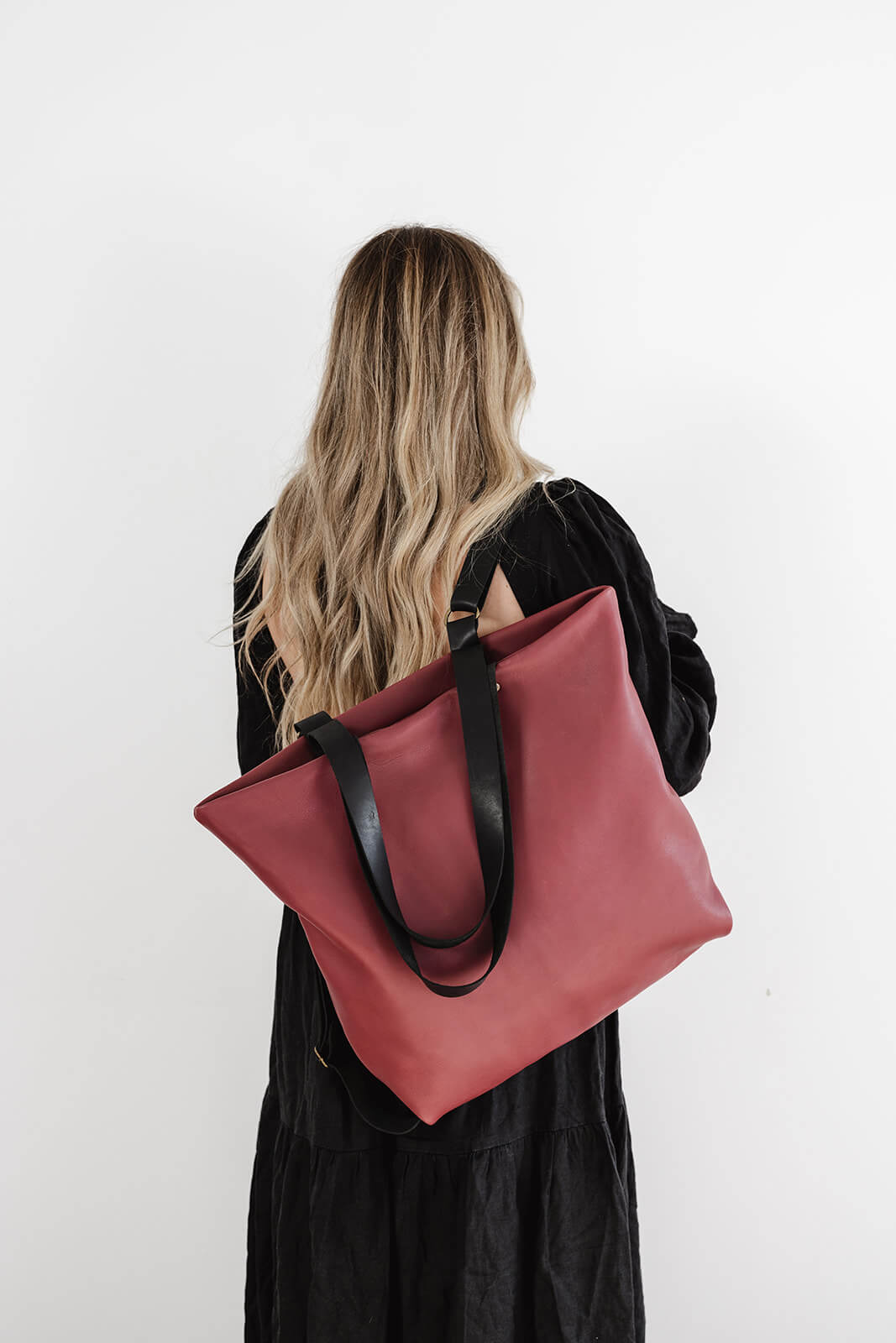 Product pic of the Ella Jackson Cherry Leather Backpack & Tote being worn as a backpack on one shoulder. The woman has long flowing blonde hair, is wearing a long black dress and is standing in front of a white background