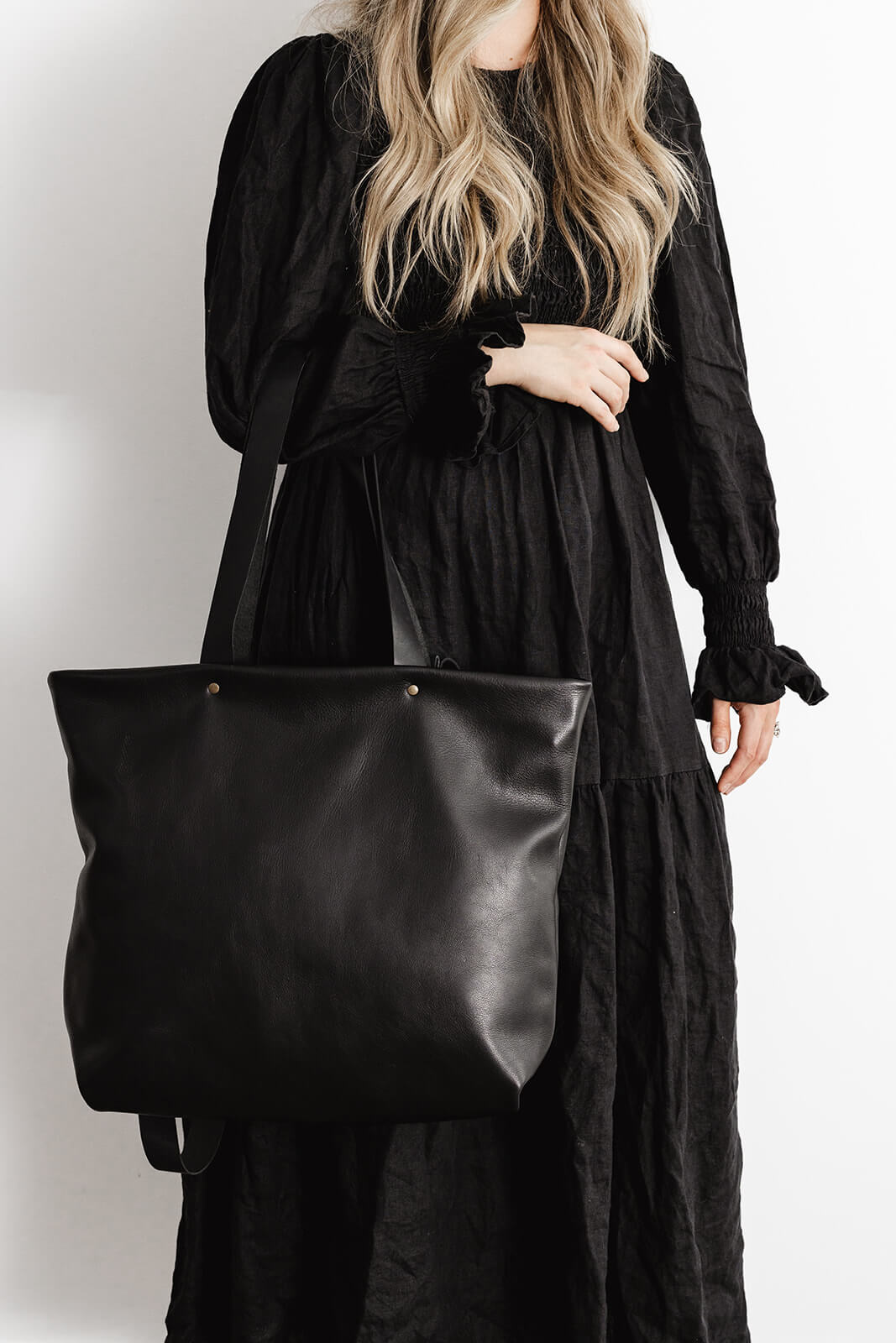 Woman with long flowing hair and wearing black dress modelling Ella Jackson Leather Backpack & Tote in Black as a tote on her arm. Photo does not show face