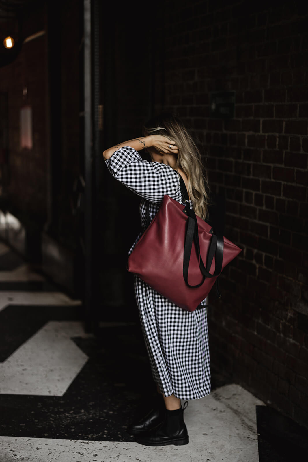 Woman standing in alley with brick wall and black and white ground. She is wearing black and white dress, black boots and a cherry leather backpack with black straps. The backpack is the Cherry Leather Backpack & Tote by Ella Jackson
