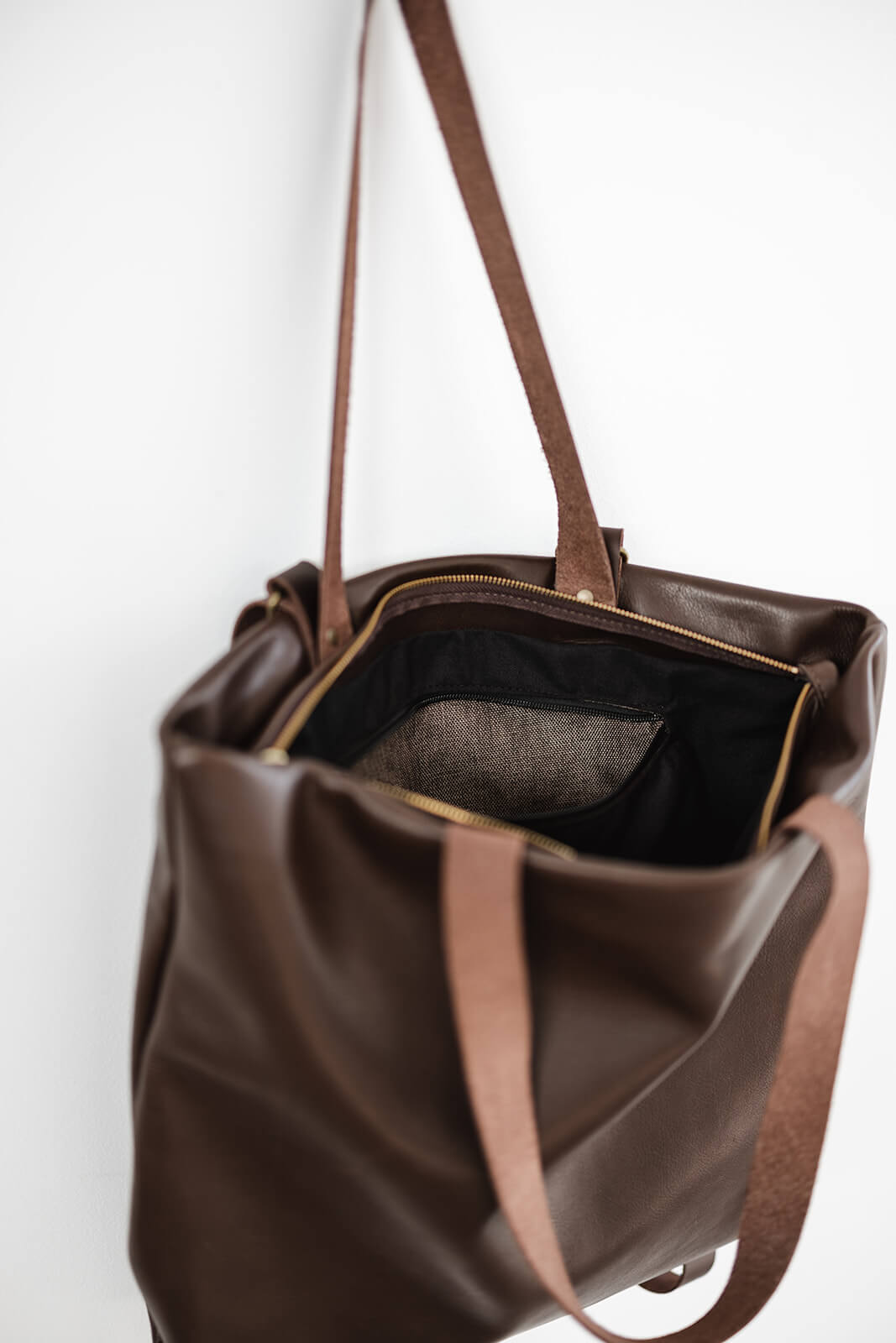Product pic of the Ella Jackson Chocolate Brown Leather Backpack & Tote. The bag is hanging as a tote on a white background. The brown and gold zip is undone to reveal the black lining and black and white fabric of the internal pocket