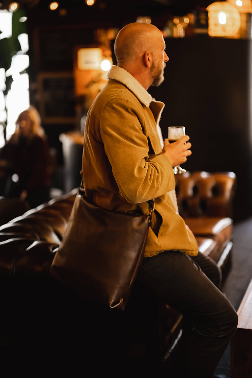 Man in sheepskin jacket sitting on arm of brown Chesterfield couch. He is holding a Guinness and wearing a brown leather crossbody bag. The bag is the Ella Jackson Chocolate Brown Leather Carryall