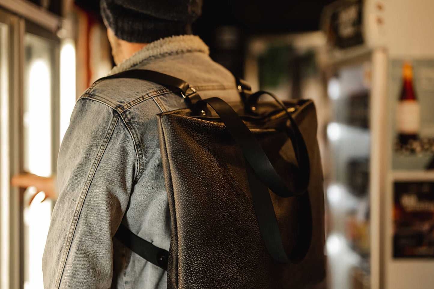 Man opening fridge in bottle shop wearing denim jacket and grey backpack with black straps. The bag is the Ella Jackson Two-tone Grey Leather Backpack & Tote