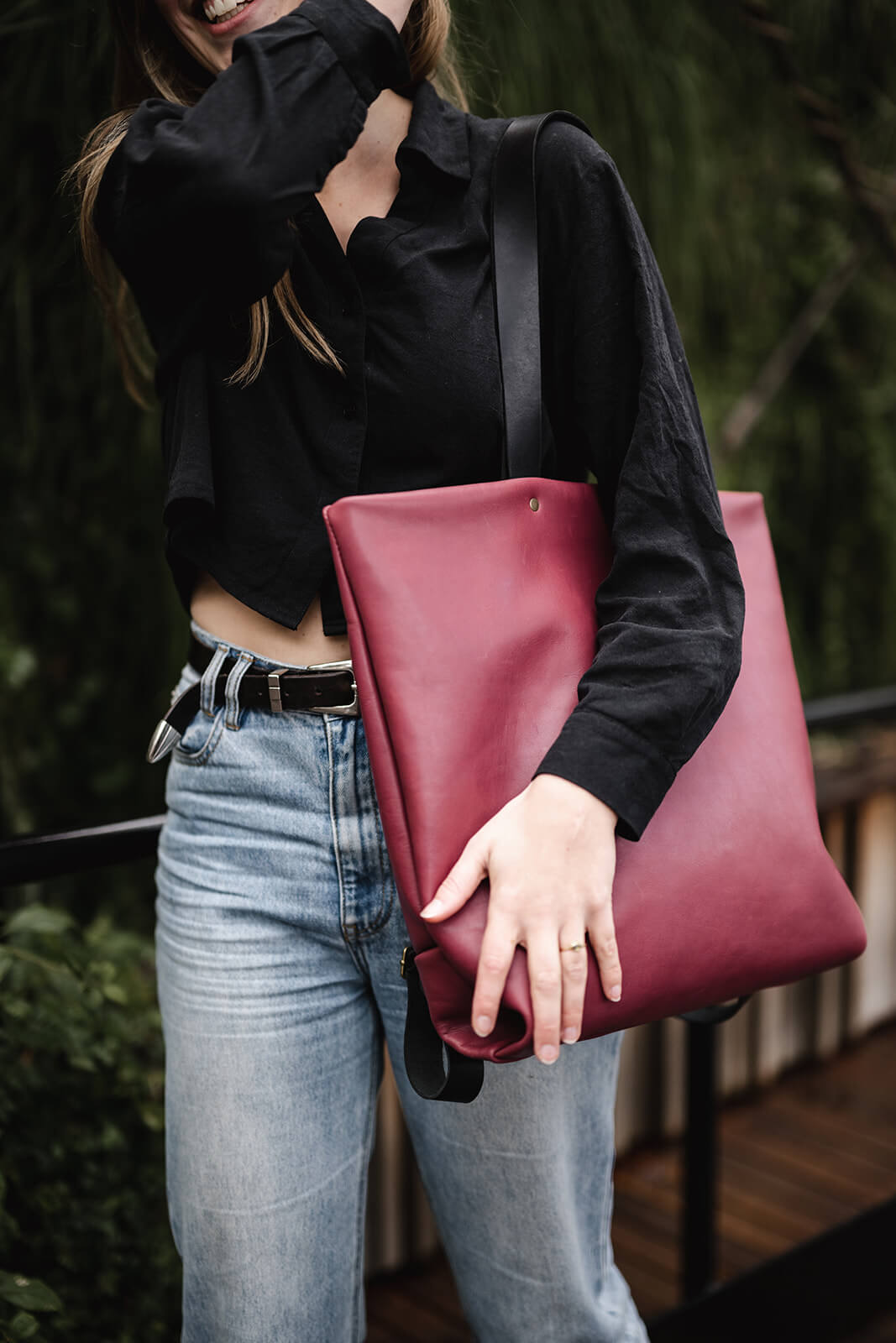 Torso photo of a woman wearing black shirt, light denim jeans and a cherry leather tote with black straps. The tote is the Cherry Leather Backpack & Tote by Ella Jackson