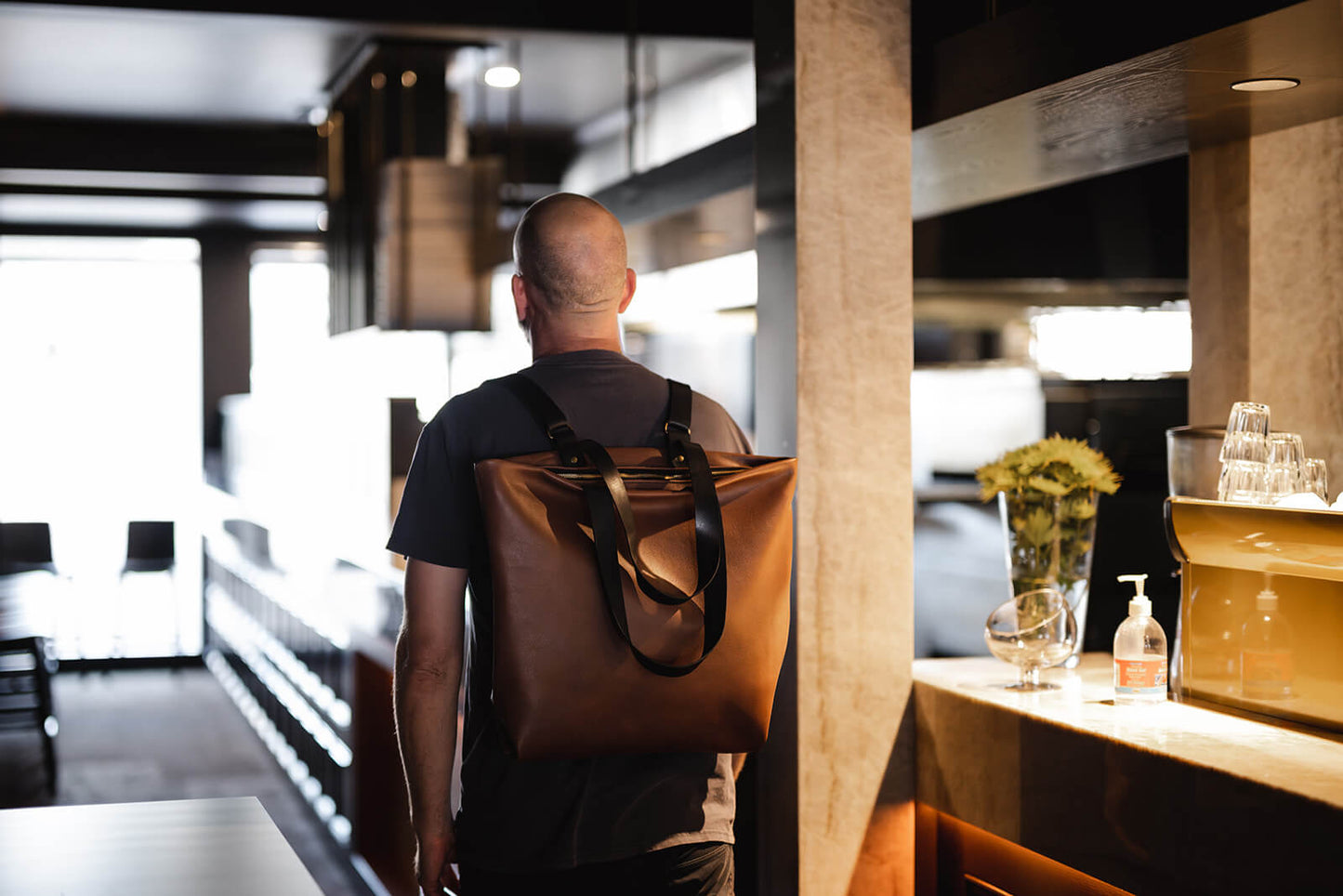 Bald man walking away from camera in a sleek restaurant and wearing a tan leather backpack with black straps. The backpack is the Tan Leather Backpack & Tote by Ella Jackson