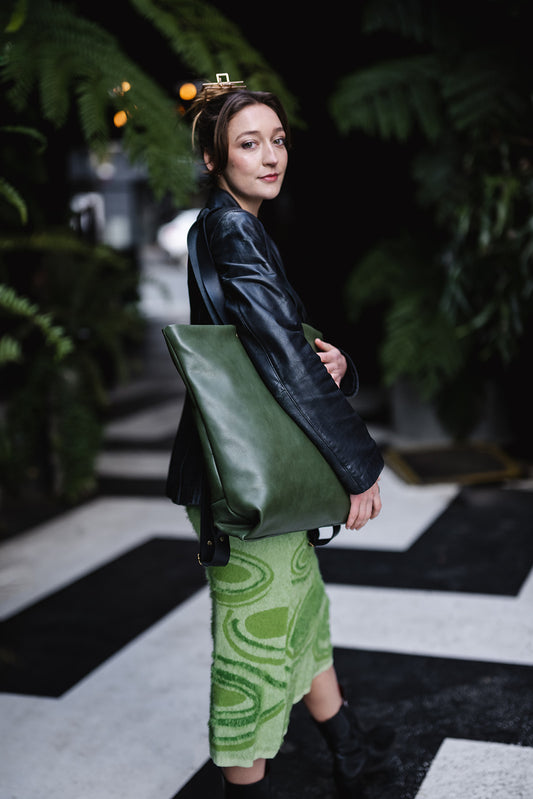 Woman standing amongst green ferns. She is wearing a green dress, black leather jacket and a green leather tote bag. The tote is the Ella Jackson Leather Backpack & Tote in Moss Green