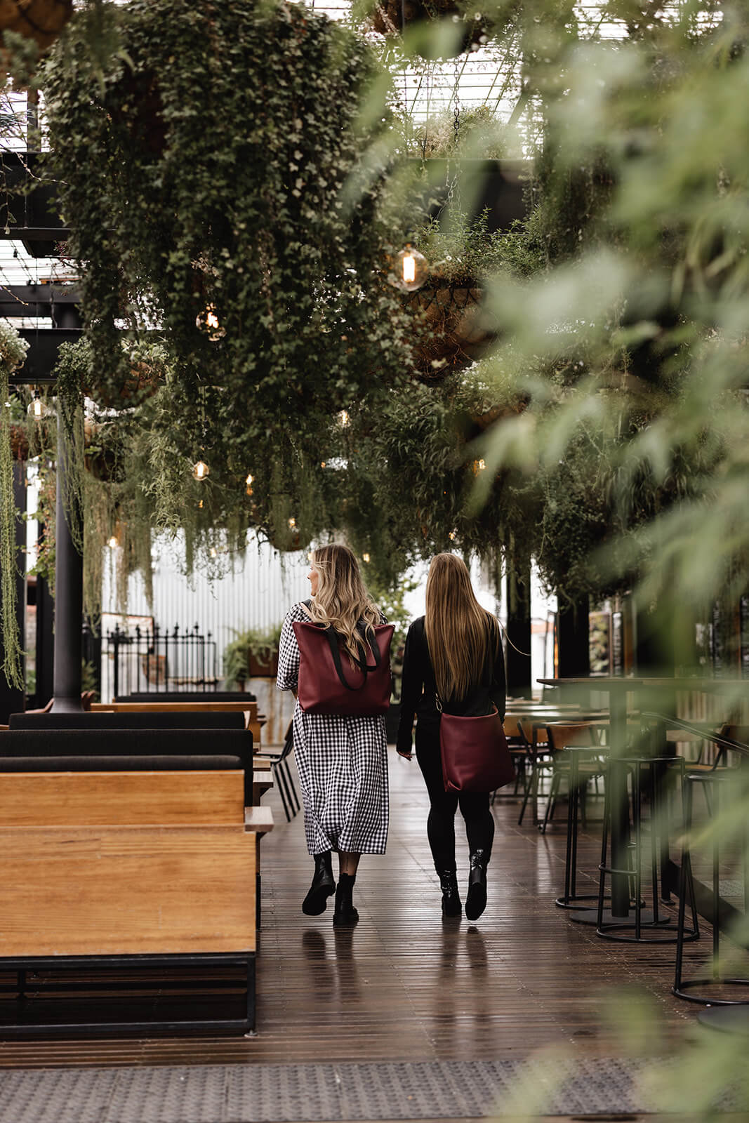 Beautiful open bar with with many hanging plants and greenery. Two woman are walking through the space wearing cherry leather bags by Ella Jackson. The woman on the left is wearing the Cherry Leather Backpack & Tote; the woman on the right is the Cherry Leather Carryall