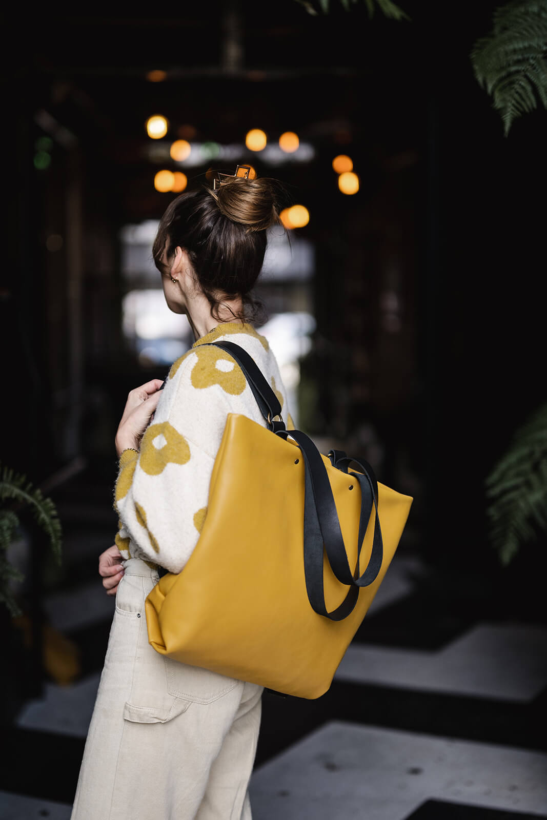Woman with dark hair up in a bun and wearing beige and yellow pants and jumper. Her back is turned and she is modelling a yellow leather backpack and tote as a backpack on her left shoulder. The straps are black leather. The bag is an Ella Jackson Yellow Leather Backpack & Tote