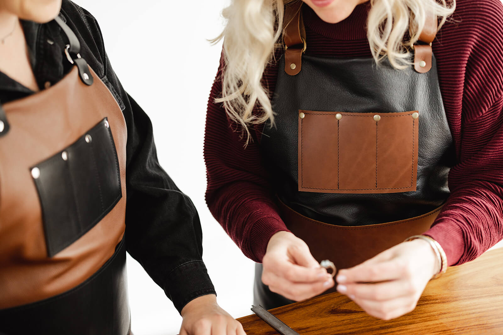 The Ella Jackson Jeweller Leather Apron being worn in 2 variations by 2 women looking at a ring. The woman on the left is wearing the variant with tan leather body, black leather straps and pockets and silver hardware. The woman on the right is wearing the variant with tan black leather body, tan leather straps and pockets and antique hardware. The bib pockets are the focus, with 3 compartments for jeweller tools