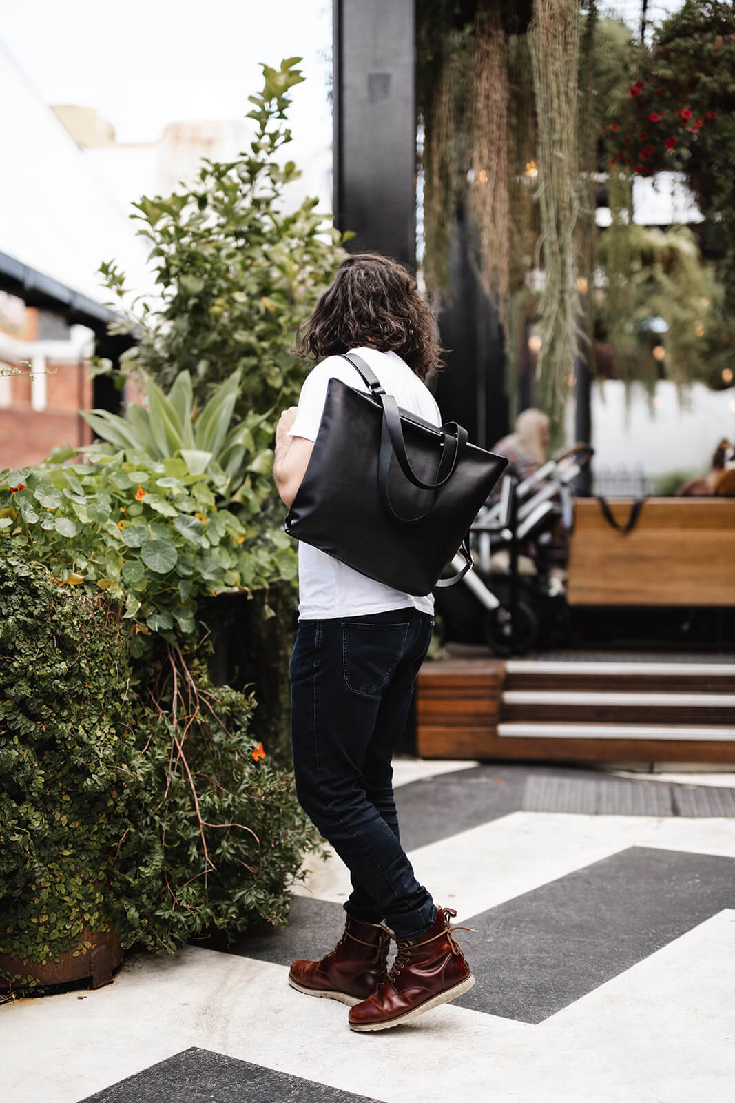 Man with long dark hair and wearing white tshirt, dark jeans and red boots modelling Ella Jackson Leather Backpack & Tote in Black. Wearing as a backpack with tote straps falling to front