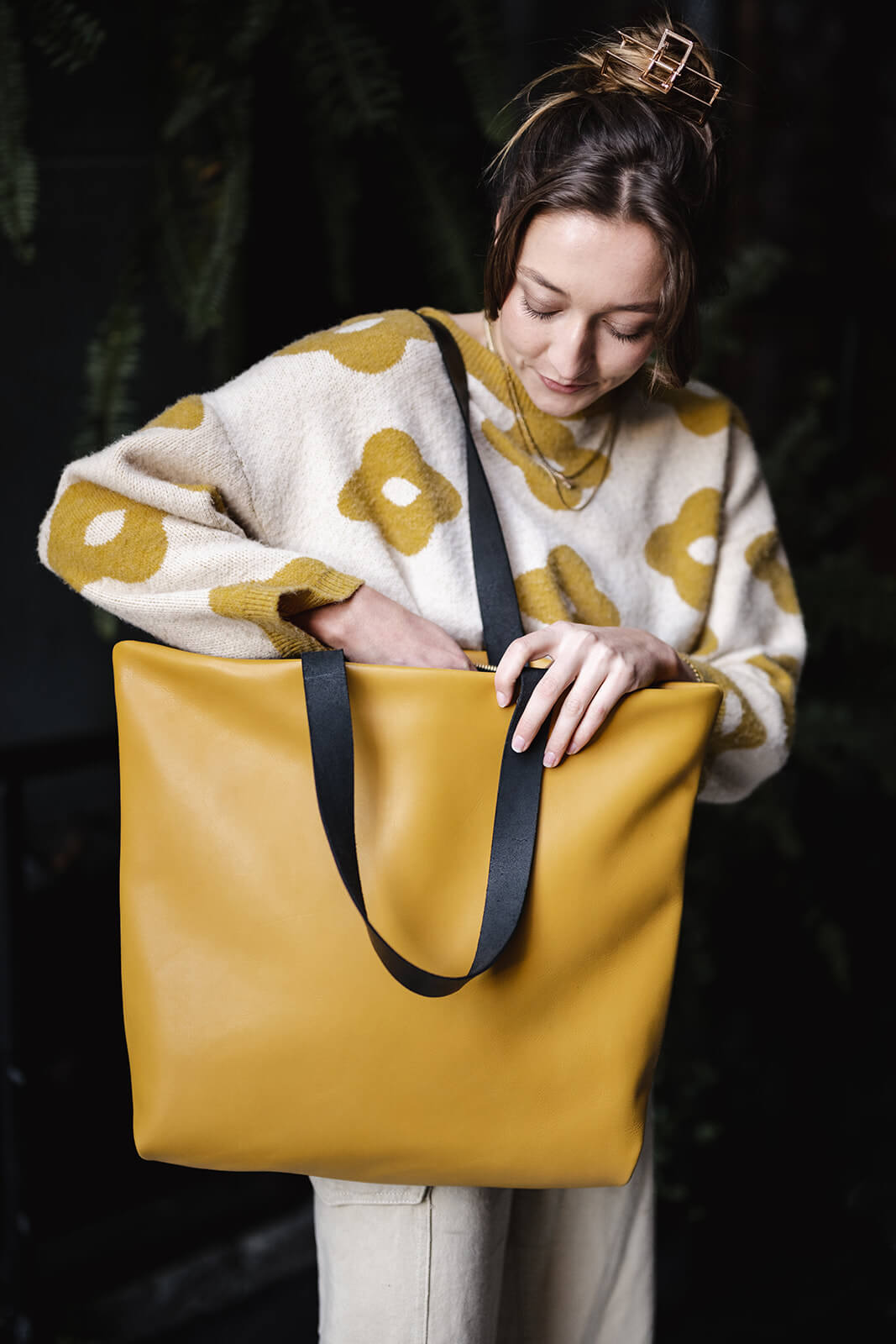 Woman wearing a yellow leather tote with black straps on her right shoulder. She is looking down into the bag with one hand searching in the bag. Her clothes are beige and yellow to complement the bag. The bag is an Ella Jackson Yellow Leather Backpack & Tote