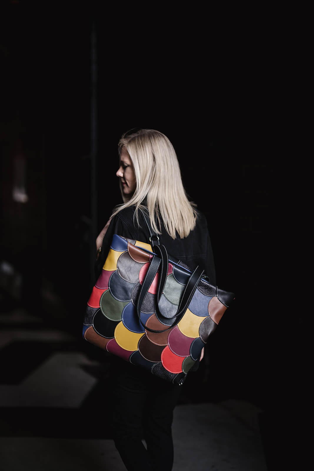 Blonde woman with black background wearing black clothes and wearing a multicoloured leather backpack on her left shoulder. The backpack is made of small leather circles in many colours stitched together. 
