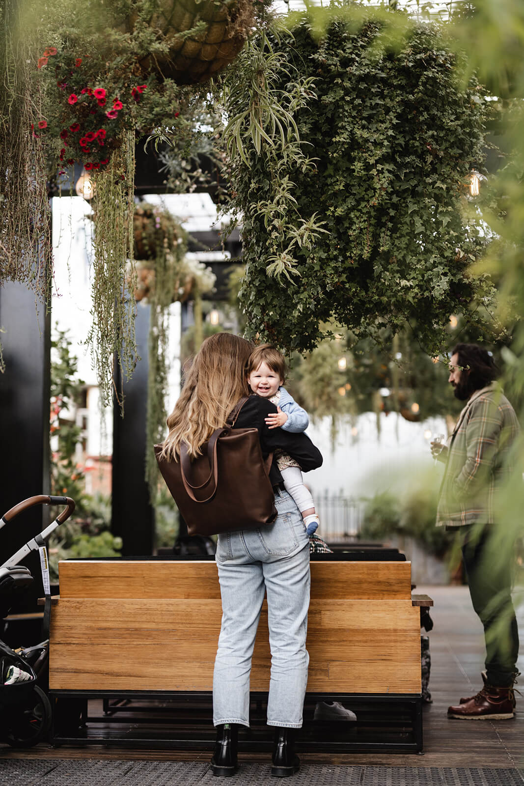 Woman with back to camera in an outdoor restaurant space with a lot of green foliage. She is holding a baby and she has a brown leather backpack on her back. The backpack is the Chocolate Brown Leather Backpack & Tote by Ella Jackson