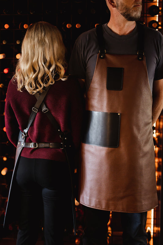 woman and man modelling leather aprons. Woman on left with back turned to show crisscross shoulder and waist black leather straps. Man on right facing forwards with tan leather apron with black pockets and straps. The aprons are the Ella Jackson Hospitality Leather Apron
