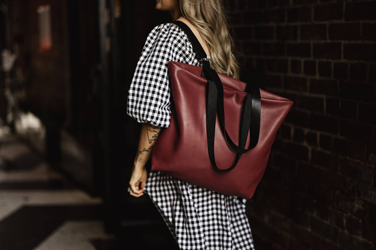 Woman in black and white gingham dress wearing a cherry leather backpack with black straps on one shoulder. The backpack is the Cherry Leather Backpack & Tote by Ella Jackson
