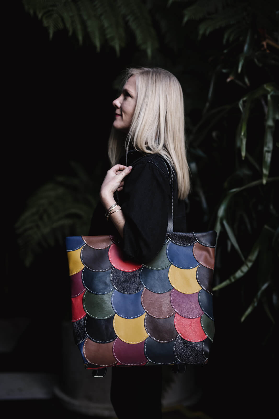 Blonde woman in black clothes standing on her side with a colourful leather tote on her shoulder. The bag is made from multiple colourful leather circles stitched together. It is the Ella Jackson Circle Bag in Multicolour