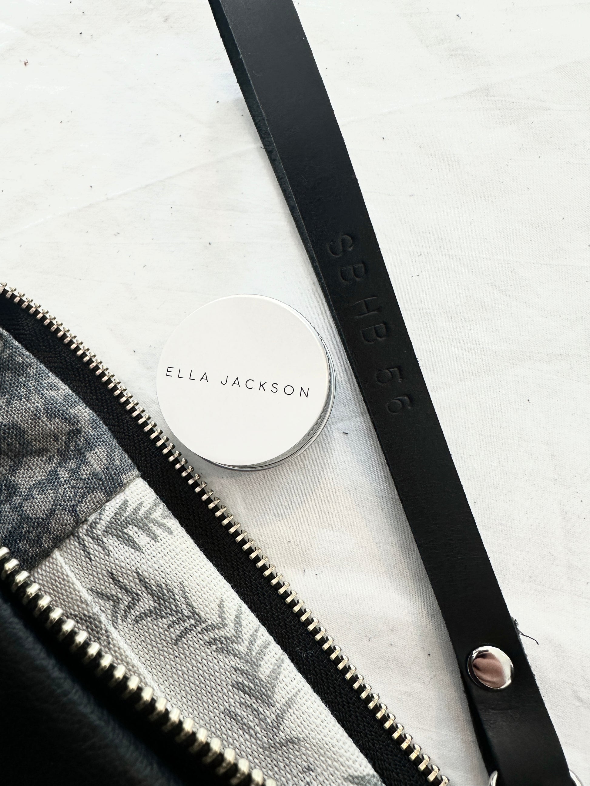 Leather clutch with detailed zipper and custom strap lettering 'SB HB 56,' next to a tin of Ella Jackson leather conditioner, placed on a light fabric background