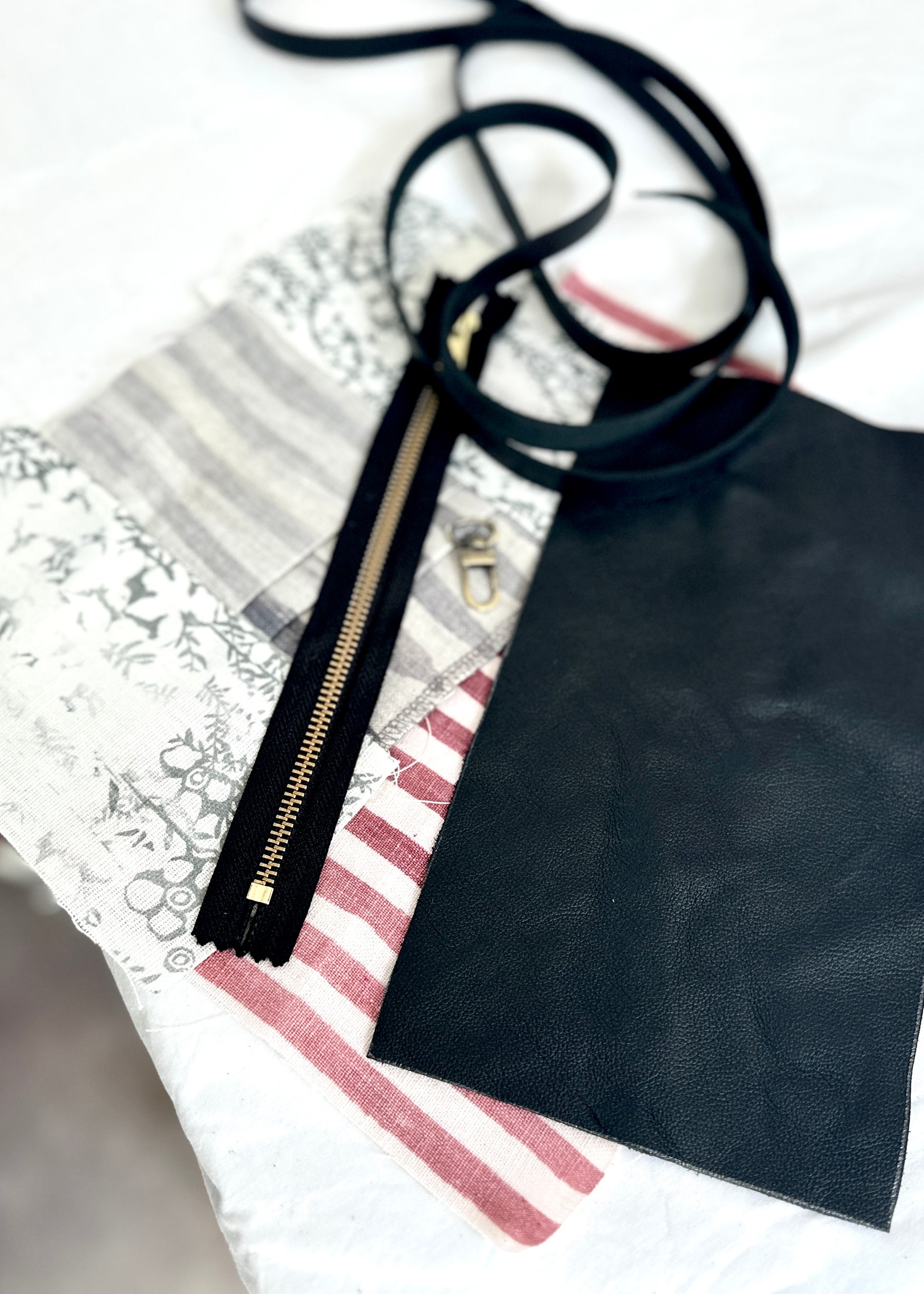 Leather clutch materials laid out, including black leather, a gold zipper, a leather strap, and a mix of striped and patterned fabric pieces, ready for assembly