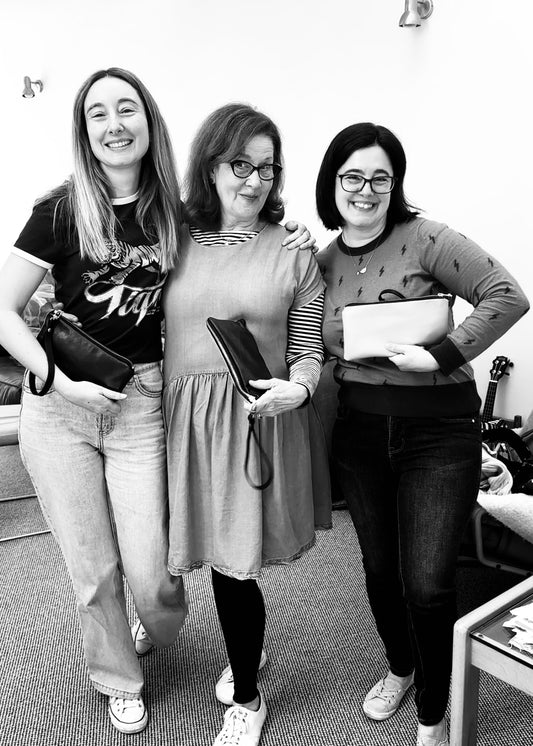 Three women smiling and proudly holding their handmade leather clutches from a workshop. The woman on the left wears a tiger-print T-shirt and jeans, the woman in the centre is in a layered dress with striped sleeves, and the woman on the right wears glasses and a patterned sweater