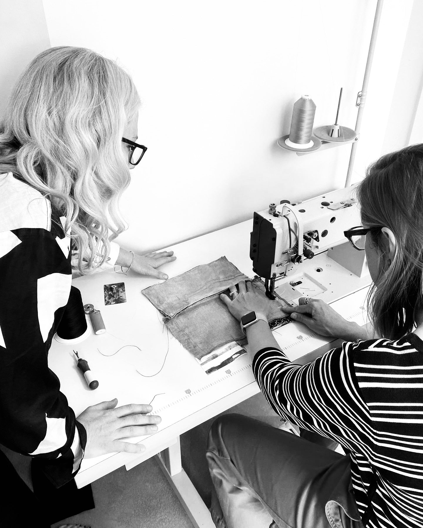 Two women working at a sewing machine during a leather workshop. One woman is guiding fabric under the machine foot while the other observes closely, both focused on the stitching process