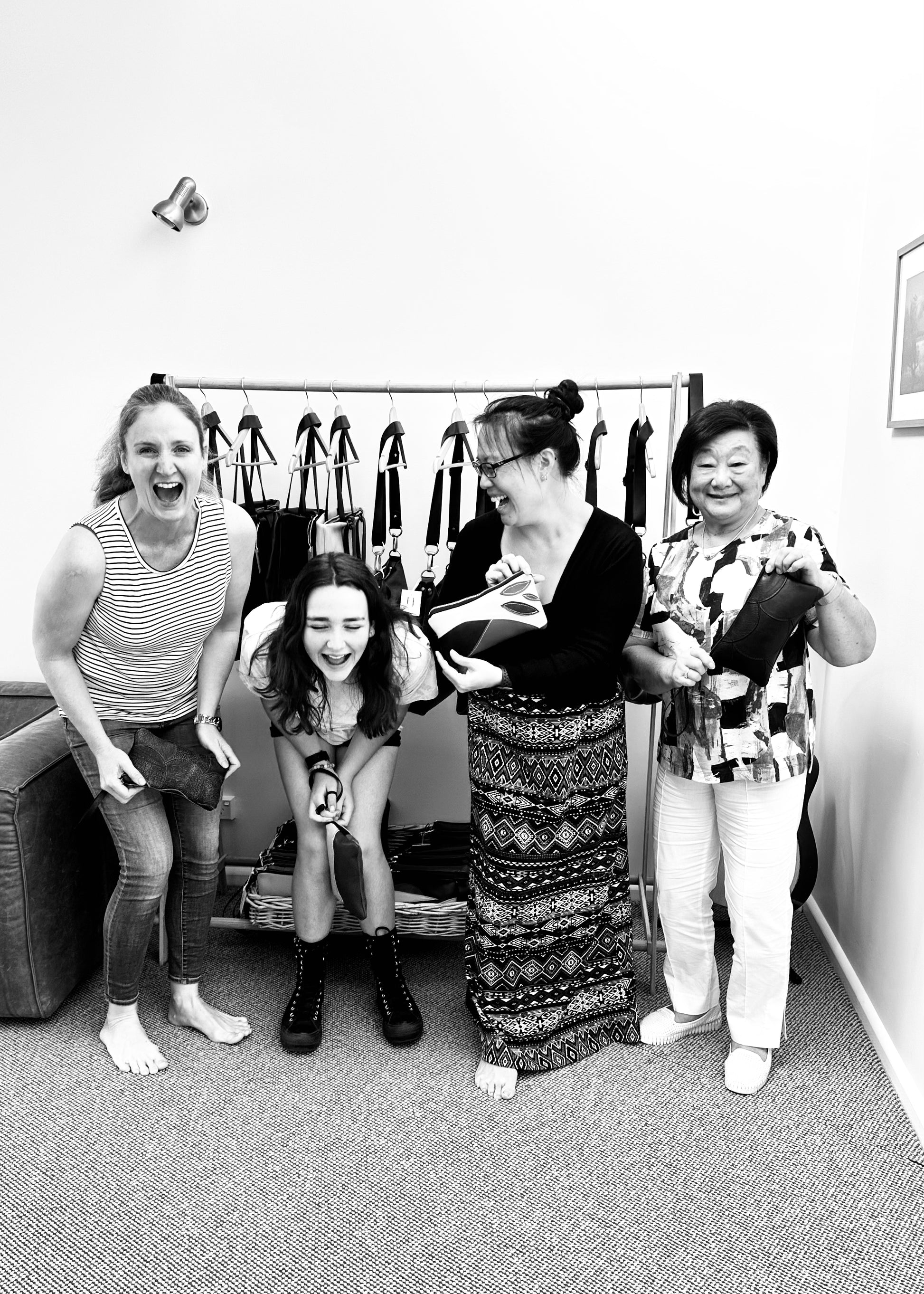 Four women laughing and holding their handmade leather clutches in a workshop setting, with a backdrop of leather handbags hanging on a rack. The group exudes joy and camaraderie as they share a fun, creative moment