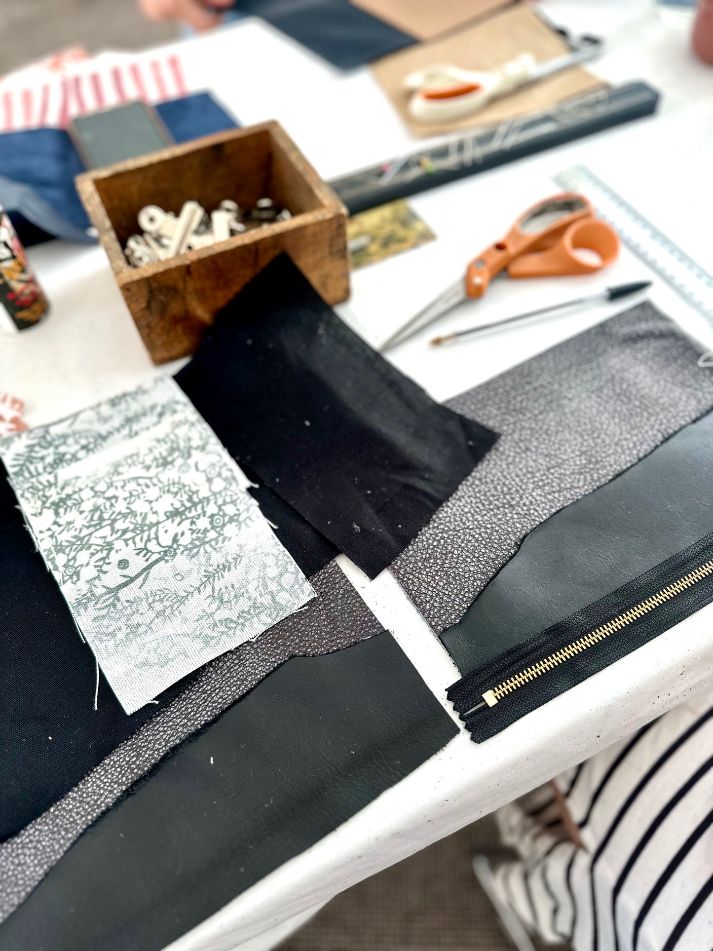 Table setup for a leather workshop featuring fabric pieces, leather swatches, a zipper, scissors, a wooden box of hardware, and various crafting tools arranged for project assembly