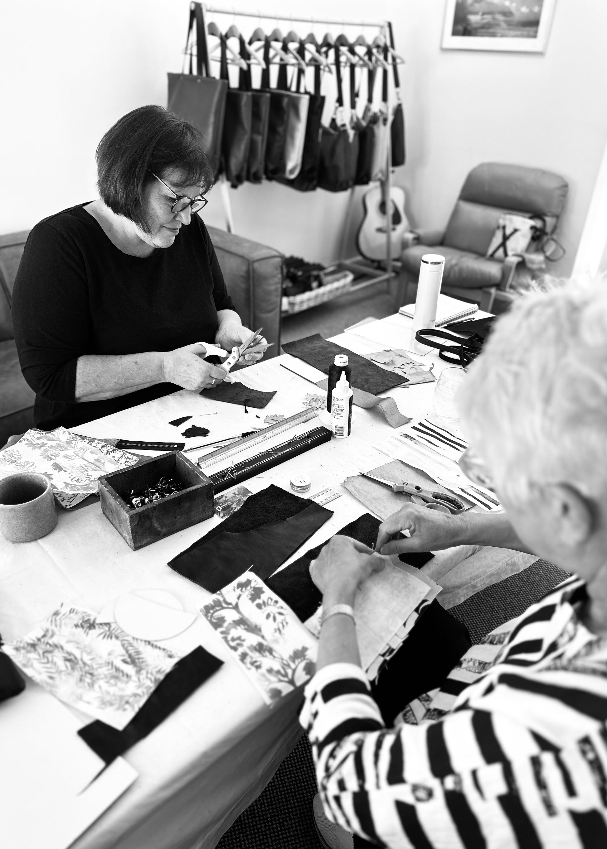 Participants working on leather projects during a workshop, surrounded by crafting materials like fabric pieces, scissors, and tools. One woman is cutting leather while another arranges fabric, with a background of finished leather handbags hanging on display